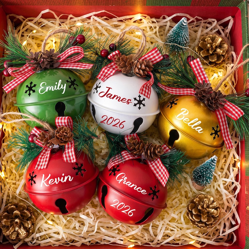 Puede incluir: Cinco adornos de cascabeles met&aacute;licos redondos en verde, blanco, dorado y rojo, personalizados con nombres como Emily, James, Bella, Kevin y Grance, decorados con lazos de cuadros rojos, agujas de pino y pi&ntilde;as peque&ntilde;as.