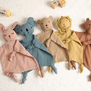 May include: A selection of baby comforters in the shape of animals, including a bunny, koala, bear, lion, and elephant. Each comforter is made from soft muslin fabric and features a knotted corner. The colours are pink, blue, beige, yellow, and brown.