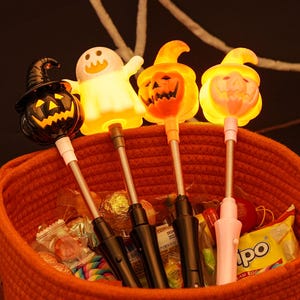 May include: An orange woven basket filled with Halloween light-up toys. The toys include a black pumpkin with a witch's hat, a white ghost, and two jack-o'-lanterns. The basket also contains wrapped sweets and treats.