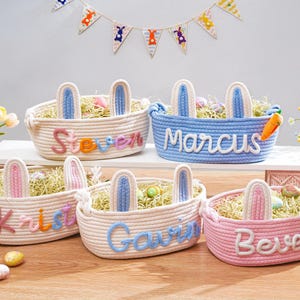 May include: Easter baskets in cream, blue, and pink, each with bunny ears and a name. The baskets are filled with decorative grass and Easter eggs. A carrot is attached to one basket. A banner with bunny shapes hangs in the background.