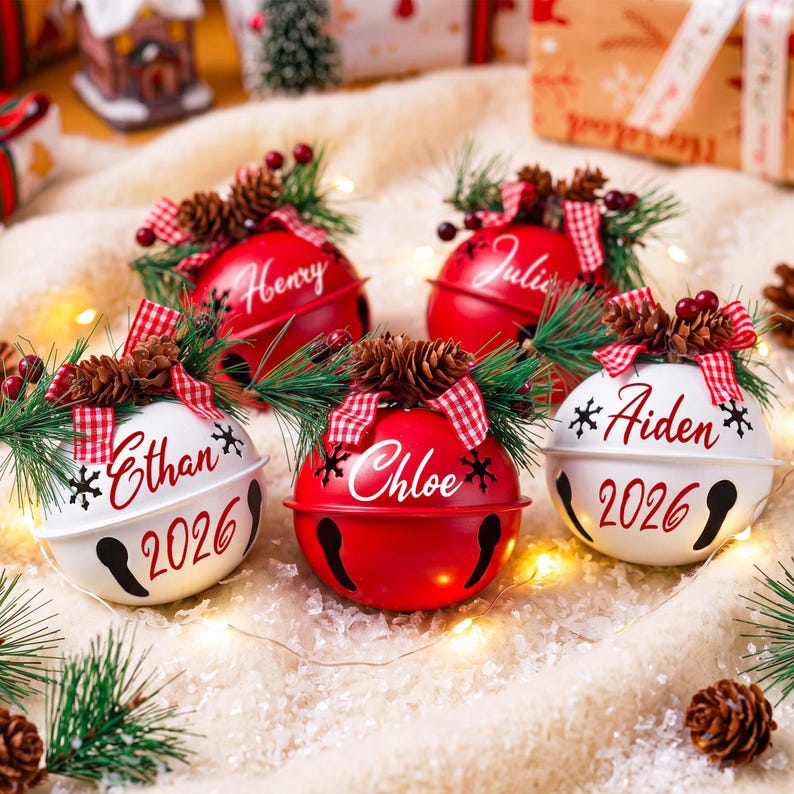 Puede incluir: Cinco adornos navide&ntilde;os de cascabeles met&aacute;licos en rojo y blanco, personalizados con un nombre y el a&ntilde;o 2026. Los adornos est&aacute;n decorados con agujas de pino, peque&ntilde;as pi&ntilde;as, bayas rojas y un lazo de cinta a cuadros rojos y blancos.