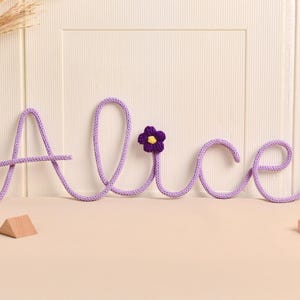 May include: A lavender-coloured, knitted name "Alice" with a purple and yellow flower accent. The letters are connected and sit on a light-coloured surface. A wooden triangle and a pink rectangular block are in the foreground.