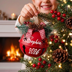 Puede incluir: Un adorno navide&ntilde;o de metal rojo en forma de cascabel con el texto blanco Sophia y 2026. El adorno est&aacute; decorado con una cinta de cuadros rojos y blancos, peque&ntilde;as pi&ntilde;as y agujas de pino verdes, colgado de un cordel r&uacute;stico.