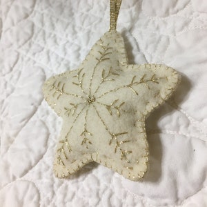 Könnte beinhalten: Cremefarbener sternförmiger Schmuck mit goldgesticktem Schneeflockenmuster und goldener Schleife. Der Filzschmuck hat goldene Nähte an den Rändern. Eine festliche Dekoration für die Feiertage.