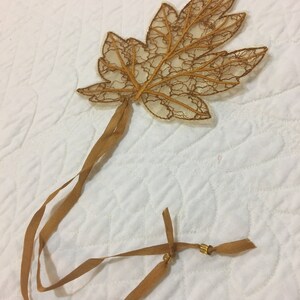 Könnte beinhalten: Dekoratives Blattornament mit filigranen Spitzenverzierungen in Gold- und Brauntönen. Das Blatt ist an einem langen, dünnen Band in Gold befestigt, das mit einer kleinen goldenen Perle endet. Geeignet für Heimdekoration oder Bastelprojekte.