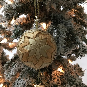 Könnte beinhalten: Ein goldfarbener Weihnachtsbaumschmuck mit Sternmuster, der an einer goldenen Kette hängt. Der Schmuck ist aus Stoff gefertigt und mit kleinen Perlen verziert. Der Hintergrund ist ein schneebedeckter Weihnachtsbaum mit Lichtern.