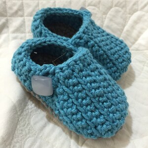 Könnte beinhalten: Ein Paar handgefertigte Baby-Schuhe in leuchtendem Türkisblau. Der Häkelstich erzeugt ein strukturiertes Aussehen. Jeder Schuh hat einen hellblauen quadratischen Knopf an der Seite. Die Innenseite ist dunkelgrau.