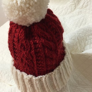 May include: A knitted winter hat with a cable knit design in red, featuring a white ribbed brim and a large white pom-pom on top. The hat is made of a thick, warm yarn.