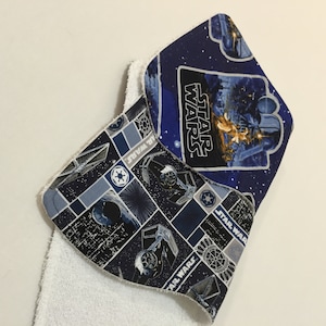 Könnte beinhalten: Zwei Baby-Lätzchen im Star Wars-Design. Eines zeigt Darth Vader und das Star Wars-Logo auf blauem Hintergrund. Das andere hat ein Muster aus Star Wars-Schiffen und Logos in Blau- und Grautönen. Beide Lätzchen haben eine weiße Frottee-Rückseite.
