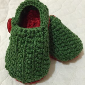 Könnte beinhalten: Handgehäkelte Baby-Booties in Grün mit rotem Innenfutter und Sohlen. Die Booties haben einen Knopfverschluss an der Seite. Das Garn hat ein strukturiertes Aussehen. Diese weichen Schuhe sind für Babys konzipiert.