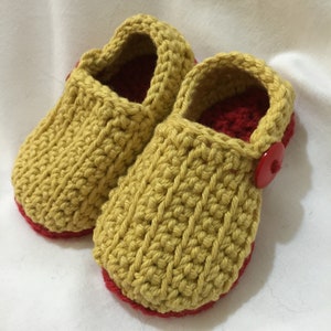 Könnte beinhalten: Ein Paar handgefertigte Baby-Häkel-Schuhe. Die Schuhe sind senfgelb mit rotem Innenfutter und Sohle. Ein roter Knopf sichert den Riemen. Die Schuhe sind aus dickem Garn gefertigt und haben ein strukturiertes Aussehen.