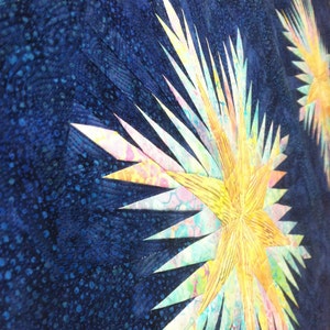 Könnte beinhalten: Nahaufnahme eines gesteppten Textils mit einem Sternmuster. Die Sternausbrüche haben gelbe, rosa und blaue Farbtöne, vor einem dunkelblauen Hintergrund mit einem gesprenkelten Muster. Der Stoff ist wahrscheinlich Baumwolle oder ein ähnliches Material.