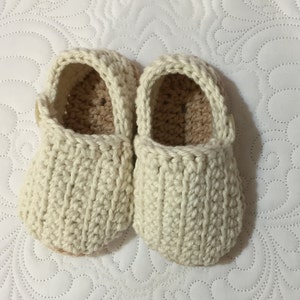 Könnte beinhalten: Ein Paar cremefarbene, handgehäkelte Babyschuhe. Die Schuhe haben eine gerippte Textur an den Seiten und eine hellbraune Innenseite. Die Oberseite jedes Schuhs hat eine kleine Schlaufe und einen Knopfverschluss. Die Schuhe werden auf einer weißen, gesteppten Oberfläche präsentiert.