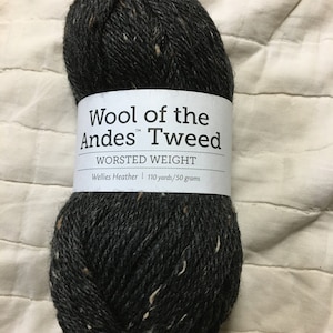 Könnte beinhalten: Ein Knäuel dunkelgrauer Wool of the Andes Tweed-Wolle mit weißen und beigen Sprenkeln. Das Etikett lautet "WORSTED WEIGHT" und "Wellies Heather 110 yards/50 Gramm."