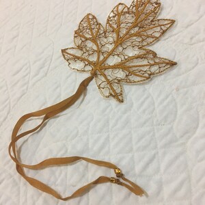 Könnte beinhalten: Ein Haaraccessoire in Form eines goldenen Ahornblattes. Das Blatt besteht aus goldfarbener Spitze mit detailliertem Adernmuster. Es hat einen braunen Stiel und passende Bänder mit kleinen goldenen Perlen an den Enden.