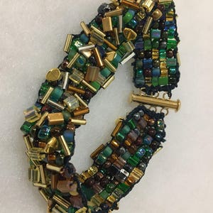 Könnte beinhalten: Ein Armband mit Perlen aus einer Mischung aus grünen, goldenen und braunen Perlen. Das Armband hat eine dunkelblaue Rückseite und einen goldfarbenen Verschluss. Die Perlen sind in verschiedenen Formen und Größen gehalten.