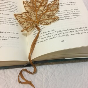Könnte beinhalten: Ein filigranes Lesezeichen in Form eines Ahornblattes. Das Blatt hat eine warme, goldbraune Farbe mit feinen Adern und Kanten. Ein passendes Band mit kleinen dekorativen Perlen erstreckt sich vom Blatt.