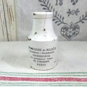 May include: A white ceramic mustard jar with a narrow neck and a wide base. The jar features black printed text that reads MOUTARDE de MAILLE Vinaigrier - Distillateur FOURNISSEUR des premières Cours DE L'EUROPE PARIS. The surface shows signs of wear.