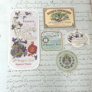 May include: A collection of five vintage-style paper labels featuring floral illustrations and text. Labels include Essence de Violettes de Parme, Essence Concentrée J. Giraud Fils, Australian Bouquet, Extrait Violette, and a circular emblem with a crest.