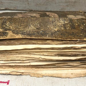 May include: An antique book with a heavily worn, dark brown cover featuring faded decorative patterns and partially legible text. The thick stack of pages is yellowed, frayed, and unevenly cut, showing significant signs of age and wear.