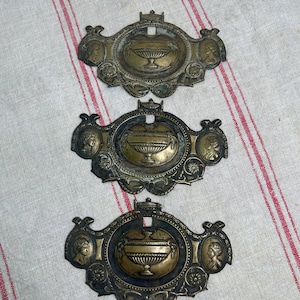 May include: Three matching brass furniture escutcheons featuring an oval central medallion with a relief of a pedestal urn. Each piece is flanked by two smaller oval portraits of figures in profile, surrounded by ornate scrollwork and floral motifs.
