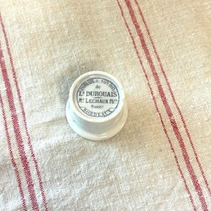 May include: A small, round, white ceramic jar lid featuring black printed text that reads: POMMADE de SAIN-BOIS, L.S DUBOUAIS, M. LECHAUX Ph.n, Sucr, BORDEAUX. The lid has a slightly raised edge and a smooth, glossy finish on its top surface.