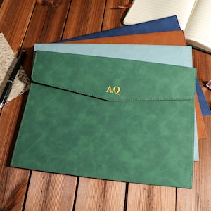 Puede incluir: Un conjunto de fundas de documentos de cuero en tonos verde, azul y marrón, sobre una superficie de madera. La funda verde superior tiene las letras doradas "AQ" en la parte delantera. También se ven un bolígrafo, un mapa y un cuaderno.