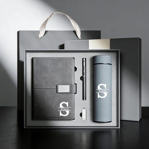Peut inclure: Un coffret cadeau gris comprenant un carnet, un stylo et une gourde. Le carnet a un fermoir argenté et la lettre "S" avec le nom "Simon" imprimé dessus. La gourde porte également la lettre "S" et le nom "Simon". L'ensemble est dans une boîte grise avec un sac cadeau gris.