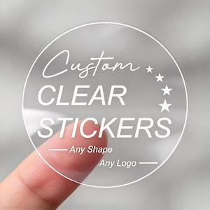 Puede incluir: Una pegatina circular transparente con texto blanco que dice Custom CLEAR STICKERS, Any Shape y Any Logo, acompañada de cuatro pequeñas estrellas blancas dispuestas en arco en el lado derecho del diseño sobre un fondo neutro.