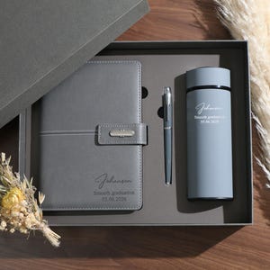 May include: A gray gift set including a notebook, pen, and tumbler, presented in a gray box. The notebook and tumbler have the name "Johansen" and the text "Smooth graduation 05.06.2026" printed on them. A small bouquet of dried flowers is included.