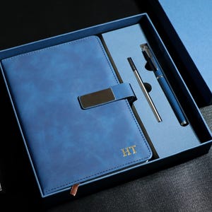 Puede incluir: Un juego de regalo azul que incluye un cuaderno con cierre metálico, un bolígrafo y una recarga de bolígrafo, todo en una caja azul a juego. El cuaderno tiene las iniciales doradas "HT" y un reloj con correa plateada al lado.