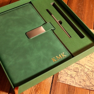 Peut inclure: Un journal en cuir vert avec un fermoir en métal argenté, présenté dans un coffret cadeau assorti. Le journal porte les initiales dorées "KMK" en relief sur la couverture. Un stylo est inclus dans la boîte, ainsi qu'une carte décorative.