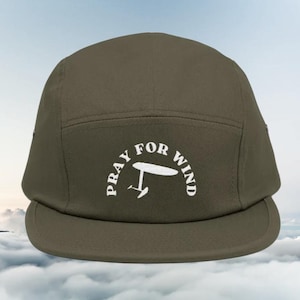 May include: Olive green five-panel cap with a flat brim. The cap features the white text "PRAY FOR WIND" in an arch, with a stylized windsurfing graphic. The background shows a cloudy sky.