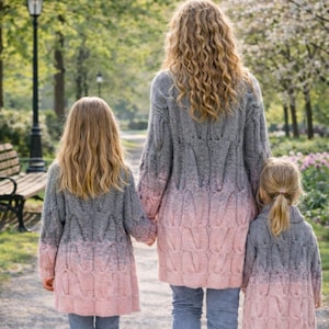 May include: Three matching cable-knit cardigans featuring a grey to pink ombre gradient. Each long-sleeved sweater has a textured pattern and is worn with blue denim jeans. The garments are shown from the back, displaying the consistent knit design.
