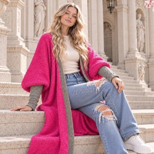May include: A long, bright pink open-front cardigan with a textured knit pattern, featuring wide sleeves and a contrasting grey ribbed trim along the edges. The garment is worn over a cream-colored top and light blue distressed denim jeans.