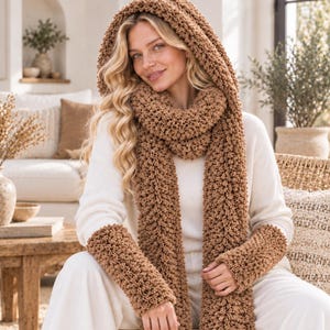 Op de afbeelding: Een set bestaande uit een bruine, gebreide sjaal met capuchon en bijpassende armwarmers. De sjaal heeft een dik, grof popcorn-breipatroon met een capuchon en lange uiteinden. De armwarmers zijn gemaakt van hetzelfde bruine garen met textuur.