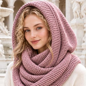 Op de afbeelding: Een oudroze capuchonsjaal met een grove ribbelsteek breitextuur. Het accessoire is ontworpen met een diepe, geïntegreerde capuchon die rond de nek en schouders valt, wat zorgt voor een dikke, gelaagde uitstraling in een zachte garenkwaliteit.