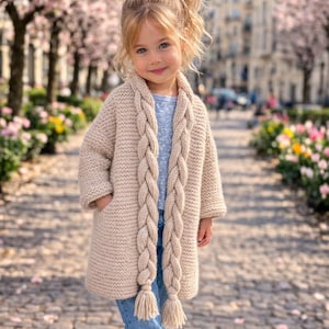 Op de afbeelding: Een jong kind draagt een beige gebreid vest met lange, gevlochten details en kwastjes. Het vest wordt gecombineerd met een blauwe spijkerbroek en een blauwe strik in het haar. De achtergrond toont een vage stadsstraat met bloeiende bomen.