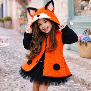 Puede incluir: Abrigo con capucha de zorro de ganchillo naranja y negro con una falda de tul negra. El abrigo presenta una capucha con forma de cara de zorro con detalles blancos y negros, mangas negras y dos acentos circulares negros en la parte delantera.
