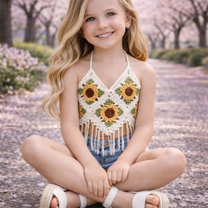 Può includere: Top all'uncinetto color crema con motivo a girasoli e frange. Indossato con pantaloncini di jeans e sandali bianchi. Lo sfondo mostra un sentiero sfocato fiancheggiato da alberi in fiore rosa.