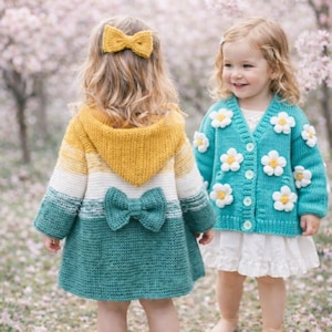 May include: Two young children wearing handmade knitwear. One child is wearing a teal and yellow hooded coat with a bow detail, the other a turquoise cardigan adorned with white daisy appliqués. Both are in a soft focus outdoor setting.