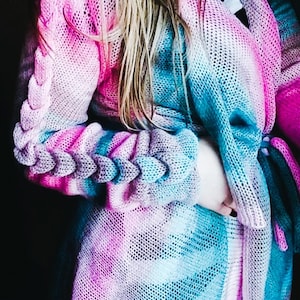 Puede incluir: Un cárdigan de punto en tonos rosa, azul y verde azulado. Las mangas presentan un diseño trenzado y el cárdigan abierto tiene un cierre de lazo. La prenda está hecha de un material ligero de tejido abierto.