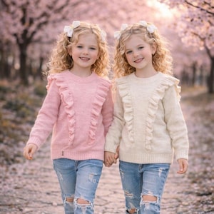 Op de afbeelding: Twee jonge meisjes dragen roze en crèmekleurige truien met ruches en een versleten blauwe jeans. Ze houden elkaars hand vast en lopen op een pad omzoomd met roze bloesem bomen. Elk meisje heeft krullend blond haar en een witte strik.