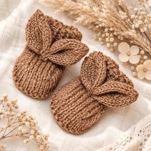 Könnte beinhalten: Ein Paar braune, gestrickte Babyschuhe mit gerippter Textur und dekorativen blattförmigen Akzenten auf der Oberseite. Die Schuhe sind aus weichem Garn gefertigt und weisen ein durchgehendes, gleichmäßiges Strickmuster in einem neutralen Farbton auf.