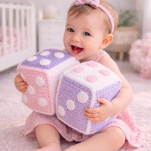 May include: A smiling baby holds two large, handmade crochet dice. One die is pink with white polka dots, the other is purple with white dots. The baby wears a pink dress and a matching headband. The dice are a soft toy for babies.