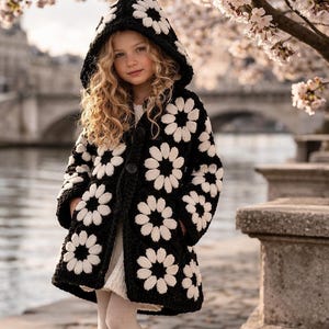 May include: A black and white crochet coat with a daisy flower pattern. The coat has a hood and is buttoned up. The person is wearing a white dress and tights. The background includes a body of water and a bridge.