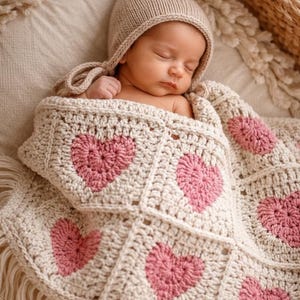 May include: A sleeping baby wrapped in a cream-colored crocheted blanket with pink heart designs. The baby wears a matching knit cap. The blanket has a fringed edge.