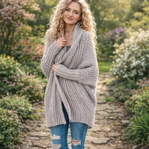 May include: A light gray, chunky knit cardigan with a shawl collar. The long-sleeved sweater is worn over a gray top and blue jeans with distressed details. The person is standing on a stone path in a garden setting.