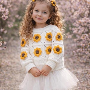 May include: A young girl wearing a cream-colored sweater adorned with yellow and brown sunflower appliqués. She also wears a white tulle skirt and a matching sunflower hair accessory. The background features soft pink blossoms.