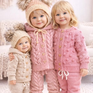 May include: Three children in matching knitted outfits. The outfits include hats with pom-poms, hooded sweaters, trousers, and booties. The outfits are in shades of pink and beige, with wooden heart-shaped buttons.